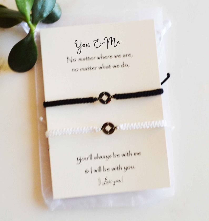 May include: A pair of matching black and white friendship bracelets with a compass charm, displayed on a card with the text "You & Me". The card also includes the phrases "No matter where we are" and "You'll always be with me".