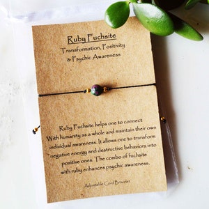 May include: A black adjustable cord bracelet with a central ruby fuchsite bead, accented by gold-colored beads. The bracelet is displayed on a brown card with text about transformation and psychic awareness. The card is in a clear plastic bag.