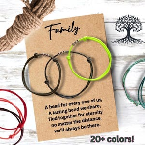 May include: Two thin cord bracelets, one black and one neon green, each featuring three small metallic beads. They are displayed on a rectangular kraft paper card with the text Family and a poem about bonds, alongside a coil of brown rope.