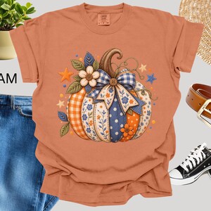 Puede incluir: Una camiseta de manga corta color naranja quemado con un gráfico central de una calabaza decorada con patrones de cuadros vichy azules y blancos, flores y lunares. La calabaza está adornada con un gran lazo de tela, flores y estrellas.