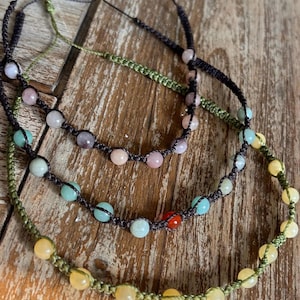 May include: Three handmade necklaces featuring gemstone beads in shades of yellow, blue, pink, and red. The beads are strung on dark brown and green cords, offering a natural, earthy look. The necklaces are presented on a wooden surface.