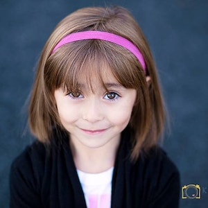 Puede incluir: Una niña pequeña con cabello castaño que lleva una diadema rosa y un cárdigan negro sonríe a la cámara.