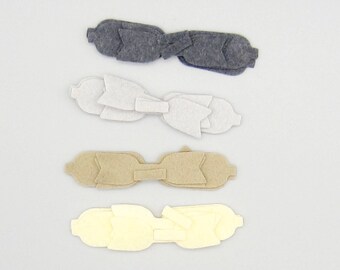 May include: Four felt hair bows in shades of gray, white, tan, and yellow. Each bow has a layered design with a central knot and rounded ends. The bows are arranged horizontally against a white background.