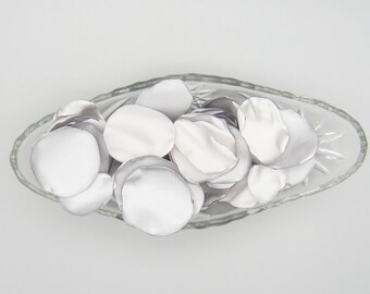 May include: A clear, oval glass dish filled with white satin flower petals. The petals are arranged in a loose pile, and the dish has a decorative pattern on the bottom. The petals are a soft, matte white, suitable for weddings or events.