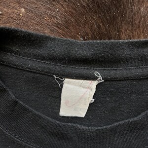 May include: Close-up of a black t-shirt collar with a white tag attached. The tag has faded pink text. The fabric of the shirt appears worn. The background is a dark brown, textured surface.