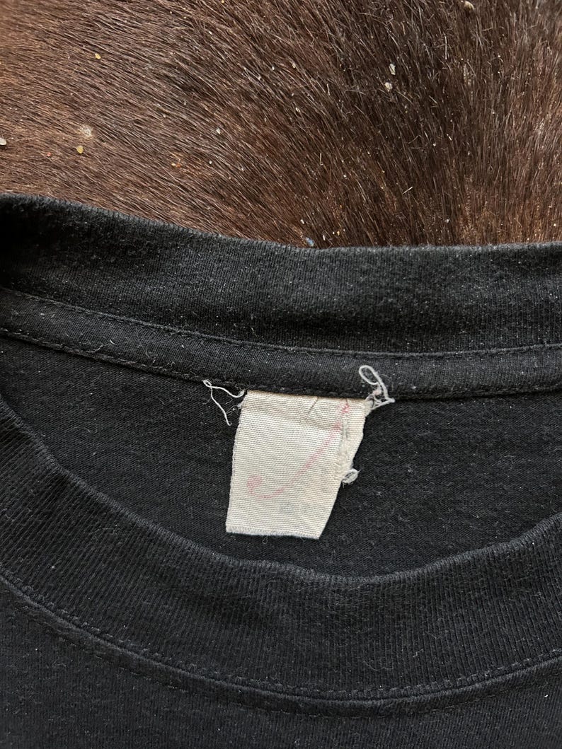 May include: Close-up of a black t-shirt collar with a white tag attached. The tag has faded pink text. The fabric of the shirt appears worn. The background is a dark brown, textured surface.