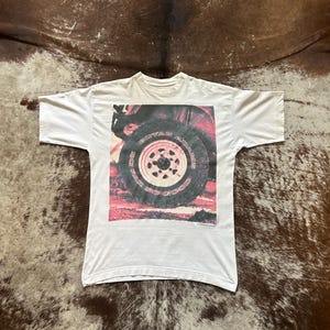 Könnte beinhalten: Weißes T-Shirt mit einem Reifengrafik und dem Text "BRYAN ADAMS" und "SO FAR SO GOOD" in Rot und Weiß. Das Shirt liegt flach auf einer braun-beigen gemusterten Oberfläche.