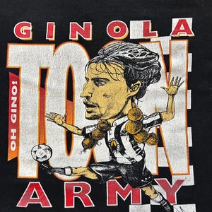 May include: Black t-shirt with a cartoon image of a footballer kicking a ball. The text "GINOLA" and "TOON ARMY" are in red and white block letters. The shirt also includes the text "OH GINOLA!" and "Copyright David James 1996".