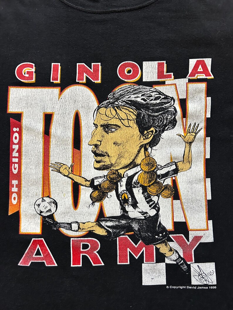 May include: Black t-shirt with a cartoon image of a footballer kicking a ball. The text "GINOLA" and "TOON ARMY" are in red and white block letters. The shirt also includes the text "OH GINOLA!" and "Copyright David James 1996".