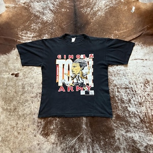 May include: Black t-shirt with a cartoon image of a footballer and the words "GINOLA ARMY" in red and white. The shirt has a crew neck and short sleeves. The design includes a football and a chequered pattern.