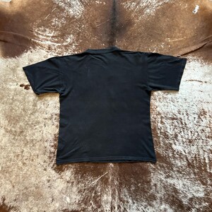May include: A black short-sleeved t-shirt, viewed from the back. The shirt is made of a soft material and has a simple design. The background is a brown and tan hide.