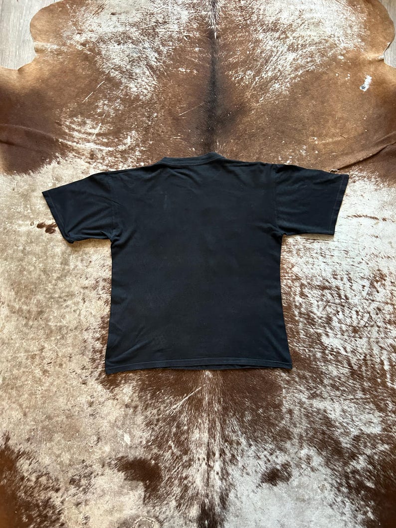 May include: A black short-sleeved t-shirt, viewed from the back. The shirt is made of a soft material and has a simple design. The background is a brown and tan hide.