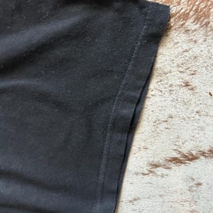 May include: Close-up of a black t-shirt sleeve. The fabric appears soft and slightly textured, with a visible seam along the edge. The background is a brown and tan patterned surface.
