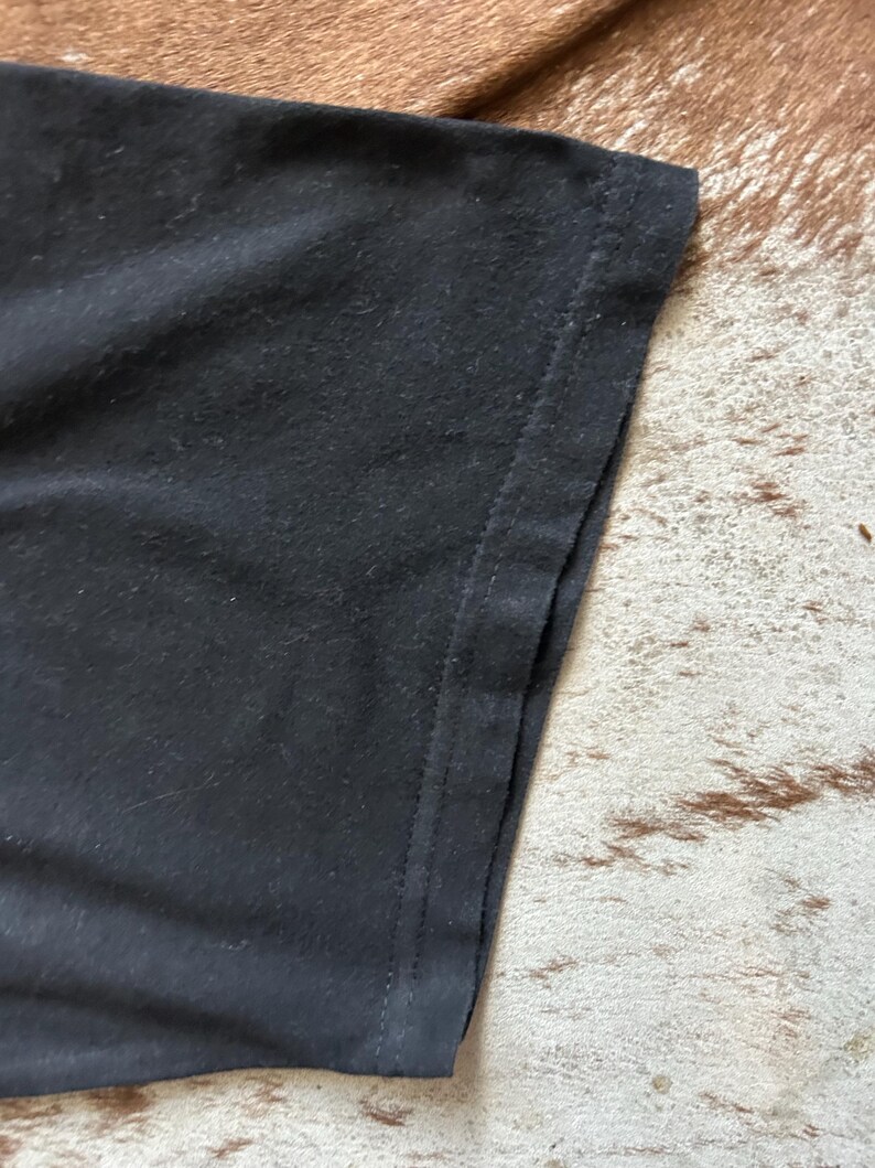 May include: Close-up of a black t-shirt sleeve. The fabric appears soft and slightly textured, with a visible seam along the edge. The background is a brown and tan patterned surface.