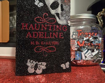 May include: A black book titled "Haunting Adeline" with a skull and butterfly design, embellished with rhinestones. The book is on a red sequined surface, next to a jar labeled "Trigger Warning".