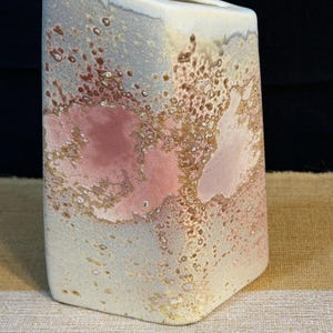 Peut inclure: Un vase en céramique avec une ouverture supérieure inclinée, présentant un émail texturé dans des tons crème, rose tendre et brun terreux. La surface affiche un motif moucheté et tacheté avec des couleurs superposées créant un aspect organique.