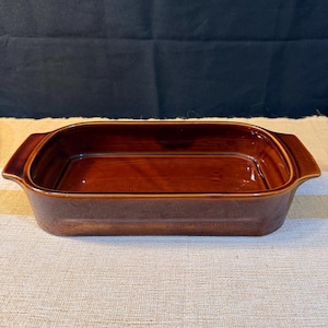 May include: A rectangular, brown ceramic baking dish with rounded corners and two handles. The dish has a glossy finish and is set on a beige and white striped surface. The background is a dark blue.