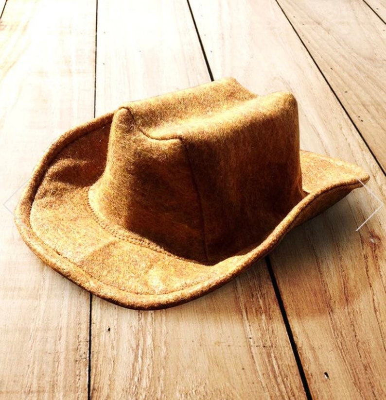 May include: A brown cowboy hat with a wide brim and a structured crown. The hat is made of a textured material and sits on a wooden surface. The hat's design is classic and functional, suitable for outdoor activities or costume wear.