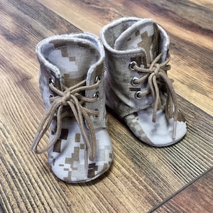 May include: A pair of baby booties in a camouflage pattern with brown laces. The booties are made of a soft fabric and have a rounded toe.