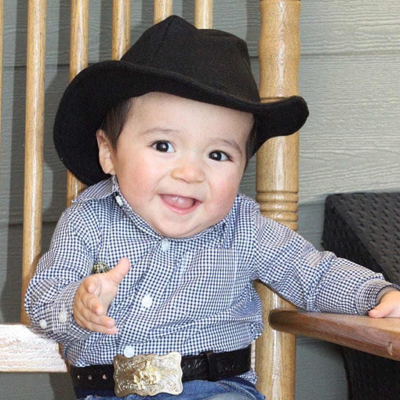 May include: A smiling baby wearing a black cowboy hat, a blue and white checkered shirt, and a belt with a decorative buckle. The baby is sitting on a wooden chair, with a happy expression.