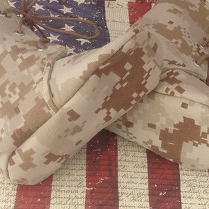 May include: A pair of tan camouflage boots with brown laces. The boots are lying on top of an American flag.