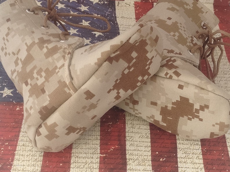 May include: A pair of tan camouflage boots with brown laces. The boots are lying on top of an American flag.