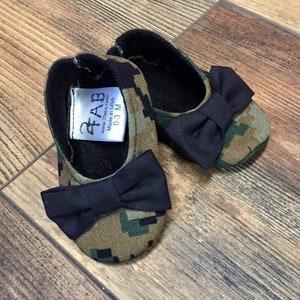 May include: A pair of camouflage baby shoes with black bows. The shoes have a label that reads "2 Babes, www.2babes.com, Made in USA, 0-3 M".