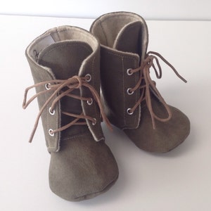 May include: A pair of olive green suede baby booties with brown laces and white lining. The booties have a lace-up closure and a rounded toe.