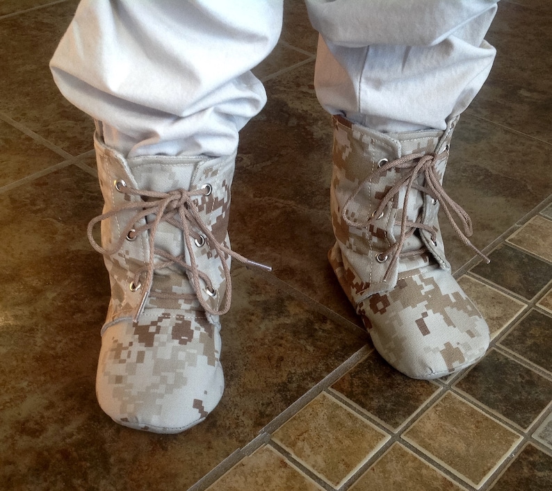 May include: A pair of tan camouflage print ankle boots with laces. The boots are made of a soft, canvas-like material and have a rounded toe.