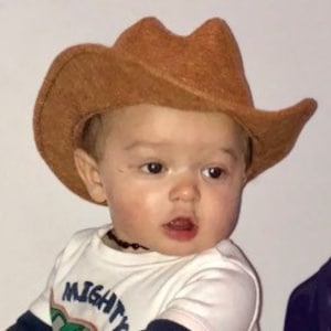 May include: A light brown cowboy hat with a wide brim. The hat is made of a textured material. The hat is worn by a baby wearing a white shirt with the text "MIGHTY".