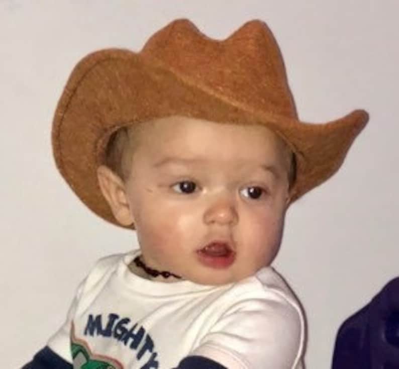 May include: A light brown cowboy hat with a wide brim. The hat is made of a textured material. The hat is worn by a baby wearing a white shirt with the text "MIGHTY".