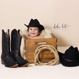 May include: A baby in a wooden crate wearing a black cowboy hat, with a pair of black cowboy boots and a coiled rope. A second black cowboy hat is on the right. The image is a western-themed baby photoshoot.