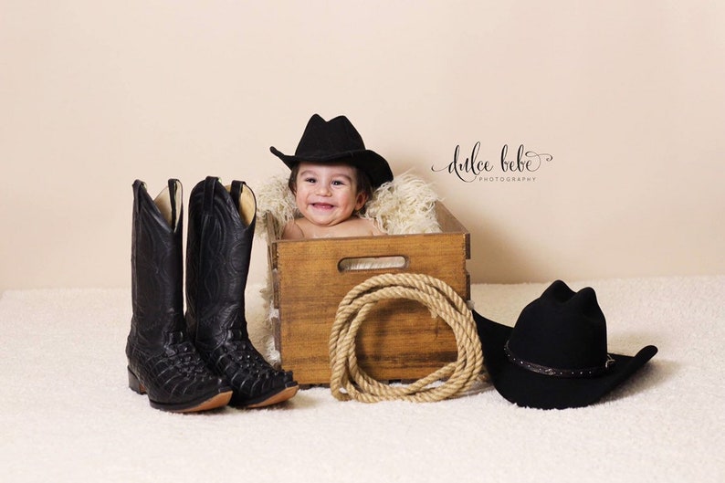 May include: A baby in a wooden crate wearing a black cowboy hat, with a pair of black cowboy boots and a coiled rope. A second black cowboy hat is on the right. The image is a western-themed baby photoshoot.