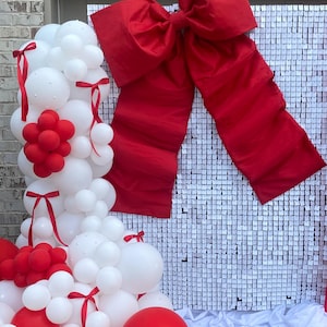 May include: A festive display featuring a large red bow and a balloon garland. The garland is composed of white and red balloons, some tied with red ribbons. A backdrop of white square sequins adds a shimmering effect.