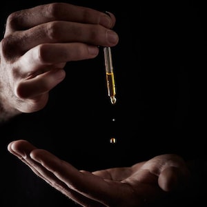 May include: A hand holds a glass dropper with a clear liquid, and a drop falls from the dropper onto an open palm.