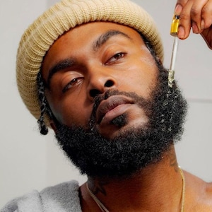 May include: A person with a beard applies a clear liquid to their beard with a dropper. The person is wearing a beige knit beanie and a gray shirt.