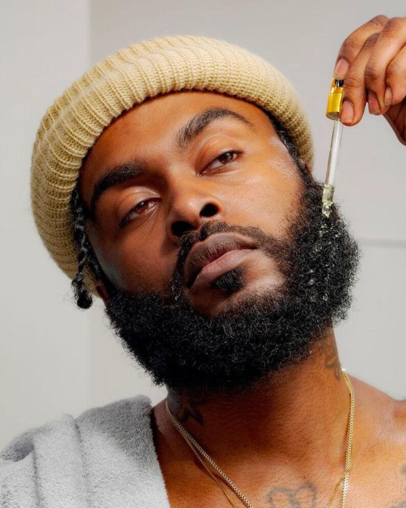 May include: A person with a beard applies a clear liquid to their beard with a dropper. The person is wearing a beige knit beanie and a gray shirt.