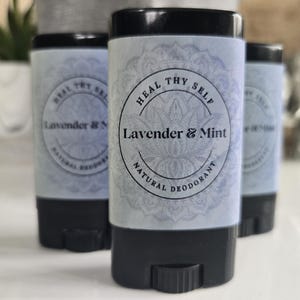 Könnte beinhalten: Drei schwarze, zylindrische Deodorant-Stifte mit hellblauen Etiketten. Die Etiketten zeigen ein kreisförmiges Mandala-Design, den Text "HEAL THY SELF" und die Worte "Lavender & Mint" in Fettdruck, gefolgt von "NATURAL DEODORANT" in kleineren Großbuchstaben.
