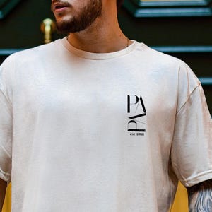 Könnte beinhalten: Beiges übergroßes T-Shirt mit kurzen Ärmeln. Das Shirt hat eine schwarze Grafik auf der Brust mit der vertikal gestapelten Aufschrift "PA" und darunter "Est. 2003". Das Shirt wird mit einer schwarzen Hose kombiniert.