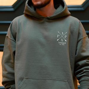 Könnte beinhalten: Ein olivgrüner Kapuzenpullover mit einer Vordertasche. Auf der Brust befindet sich ein weißes Logo mit den Buchstaben "P" und "A", gekreuzten Rudern und dem Namen "Leonard". Der Pullover ist aus weichem Material und hat eine Kapuze.