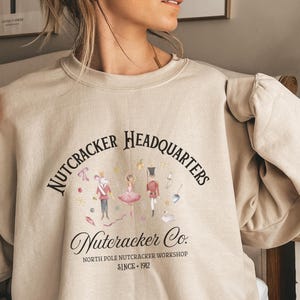 Könnte beinhalten: Beiges Sweatshirt mit dem Text "Nutcracker Headquarters" und einer Grafik von Nussknacker-Charakteren. Das Sweatshirt trägt auch den Text "Nutcracker Co. North Pole Nutcracker Workshop Since 1912".