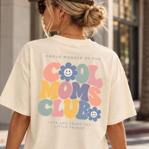 Könnte beinhalten: Ein cremefarbenes Kurzarm-T-Shirt mit einem Rückendruck, der den Text PROUD MEMBER OF THE COOL MOMS CLUB und LOVE AND ENJOY THE LITTLE THINGS in bunter Retro-Typografie sowie zwei blaue Blumen-Icons mit lächelnden Gesichtern zeigt.