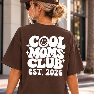 Könnte beinhalten: Ein dunkelbraunes T-Shirt mit kurzen Ärmeln und weißem Aufdruck auf der Rückseite mit der Aufschrift COOL MOMS CLUB EST. 2026, wobei ein Smiley das O in COOL ersetzt und vierzackige Sterne die fetten, geschwungenen Buchstaben umgeben.