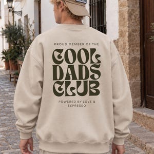 Könnte beinhalten: Ein beigefarbenes Sweatshirt mit Rundhalsausschnitt und dunkelgrünem Text auf der Rückseite, der PROUD MEMBER OF THE COOL DADS CLUB POWERED BY LOVE & ESPRESSO lautet. Das Sweatshirt wird mit einer beigen Baseballkappe mit blau-weißem Aufnäher getragen.