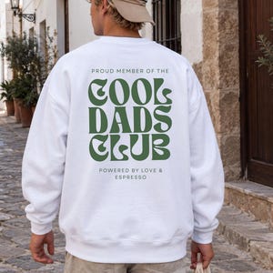 Könnte beinhalten: Ein weißes Langarm-Sweatshirt mit dunkelgrünem Text auf der Rückseite, der PROUD MEMBER OF THE COOL DADS CLUB lautet, sowie dem zusätzlichen Text POWERED BY LOVE & ESPRESSO in kleinerer Schrift unter dem grafischen Hauptdesign.