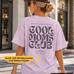Könnte beinhalten: Ein fliederfarbenes, meliertes T-Shirt mit dunkelgrauem Text auf der Rückseite, der PROUD MEMBER OF THE COOL MOMS CLUB lautet, sowie dem Namen LEONARD und EST. 2026 in einer einfachen Schriftart darunter. Das T-Shirt hat kurze Ärmel.