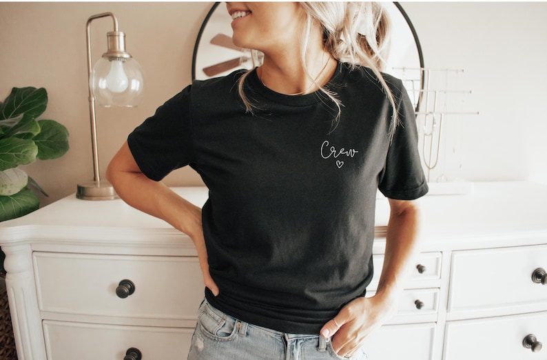 May include: Black crew neck t-shirt with the word "Crew" in cursive script and a small heart. The shirt is made of a soft material and is a solid black color. The model is wearing blue jeans.