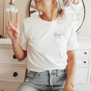 May include: White t-shirt with the word "Braut" in script and a small heart. The shirt is paired with blue jeans. The person is giving a peace sign.
