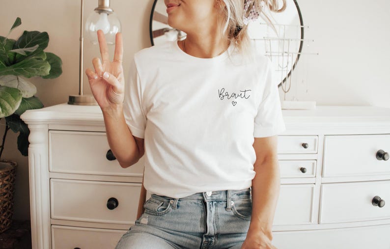 May include: White t-shirt with the word "Braut" in script and a small heart. The shirt is paired with blue jeans. The person is giving a peace sign.