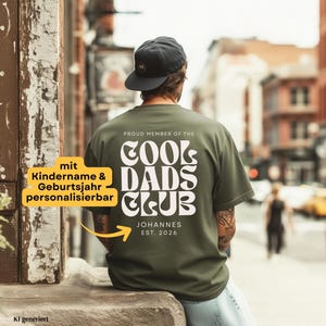 Könnte beinhalten: Ein olivgrünes T-Shirt mit weißem Text auf der Rückseite, der PROUD MEMBER OF THE COOL DADS CLUB lautet, mit dem Namen JOHANNES und EST. 2026 darunter. Das T-Shirt wird mit einer dunkelgrauen Baseballkappe getragen, Ansicht von hinten.